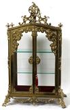 Lot# 1011 GILT BRONZE TABLETOP CURIO VITRINE, C. 1900, H 34", W 22", D 12", Glass side and door panels. Figural putto and scrolled foliate crest. Raised on scrolled bracket feet. Good quality construction. Two interior glass shelves.
