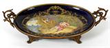 Lot# 1208 SÈVRES HAND PAINTED PORCELAIN CENTERPIECE, SIGNED, L 15", D 10", The signed Sèvres hand painted porcelain has a cobalt blue border with an oval painting of a young woman reclining at the shore of a lake. Signed M. Humez. Overall length including the double handled French gilt bronze frame is 20".