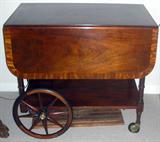Lot# 1430 MAHOGANY TEA CART, C. 1940, H 29", L 32", W 20". Satinwood inlay. Separate wood frame tray inset with glass. Drop leaves. Wood wheels. Handle is hinged for elevation.