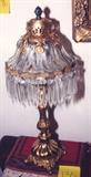 231/135  Lamp.  An early 20th century table lamp in the Victorian manner.  A molded glass shade with gilded metal mounts, the rim supporting drop prisms and faceted beads.  This raised on a gilt metal embossed post over a gadrooned dome and set on an embossed, footed base.
23 1/2" high.  $650.00
