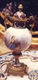 85/273  Vase.  A 19th century  French signed porcelain vase of globular form, hand painted showing a female figure in a landscape.  An ormolu finial caps the champlevé lid, scrolling handles.  The vase is raised on a champlevé stem set on a footed ormolu base.
Signed:  Valire [no listing found]
7" high:  $450.00