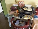 Small White Desk, Computer Printer, Elvis Singing Heart, Camera in Black Case, Camera Attachments in Burgundy Bag, Paints for Children, Desk Tray with Stationery, Variety of Colored Paper, Small Cords, White Binders, Digital Frame on Chair, Desk Chair with Cushion. Artwork, Basket of Stamp Art. Small Albums.