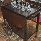 Multiple crystal and glass candlesticks sitting atop an antique two-tiered tea cart with drop leaf sides.  Removable tray needs a fresh piece of glass.