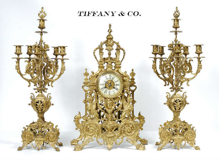 Lot 1001:  TIFFANY & CO. LOUIS XIV STYLE GILT BRONZE CLOCK GARNITURE SET: A fantastic clock garniture set by Tiffany & Co. to include gilt bronze mantle clock with applied decoration to include urn, griffon, Bacchus, masqueron, foliate and architectural motifs. Porcelain dial with Roman numerals marked Tiffany & Co., New York. Time and strike movement marked Tiffany & Compe, New-York. Serial number 3977, pendulum and back bezel with matching numbers. 25 1/4'' h. x 16 1/4'' x 6 1/2''. Sold with matching 6 light candelabra garniture set, marked 132 inside base. Measures 28 1/2'' h. x 11 1/2'' dia. Est. $6000/8000**
CONDITION: Currently running however not guaranteed to continue. Key and pendulum in office.