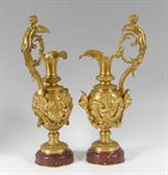 Lot 1002:  PAIR OF FRENCH DORE VICTOR PAILLARD FIGURAL BRONZE EWERS: Winged Nude Maiden Handles, relief bearded man's face on bottom side of spout, hairy dolphin masquerons with scaly tails intertwined, resting atop oxblood marble plinths, 26 3/4'' high, incised signature.