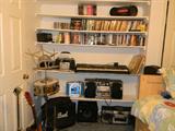 several boom boxes, drum, guitar and music stands, keyboard, CDs, lots of DVDs, VHS tapes, and more