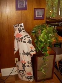 Fabric sculpture of a woman with pottery accents, next to a speaker with a plant on it