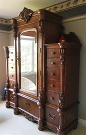 Lot#25, MODERN CARVED WALNUT, WARDROBE H 90.5", L 72", D 24"Having a carved crest and crown moldings above a center mirrored glass panel door flanked by half round fluted columns. Door opens to two shelves and two open linen drawers at the bottom. Six stacked drawers on each side of the door and one drawer below the door. Matching half round fluted columns at the front corners. All on bun feet.