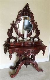 Lot#30, CARVED MAHOGANY BAROQUE VANITY, H 72", L 48", D 20"Having a shield form mirror with a pierced design frame of carved leaves, vines and grapes. All attached to a base with three narrow jewelry drawers with contoured facades. Vanity top has a contoured edge and pierced design contoured skirt with carvings of acanthus leaves with hanging finials at the corners. On four carved scrolling legs emanating from a turned spindle center post with a turned finial at the bottom.