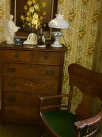 A glowing oil still life of yellow roses sets off the warm tones in this oak tall dresser. A porcelain mantle clock and a figurine with a goat are placed on top, next to a converted lantern. Check out the green leather seated armchair -- its unusually roomy.