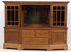 Lot#109, MISSION STYLE OAK CABINET H 60", W 82", D 18"Having a crown molding above two glass and wood framed doors flanking a center bevel glass mirror. Bottom section is slightly angled rearward at the sides and has two cabinet doors flanking three drawers, two above and one single drawer at the bottom. All with brass pulls. Flush façade across the bottom with bracket feet.