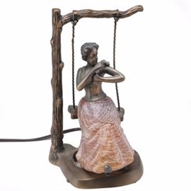Bronze Tone Table Lamp of Woman and Butterfly: A bronze tone table lamp of a woman and butterfly. This table lamp depicts a seated woman on a tree swing and a butterfly at her shoulder. The woman’s orange glass dress acts as the lamp’s shade, and the base and frame are bronze tone metal. The underside of the base is marked “Made in China.”