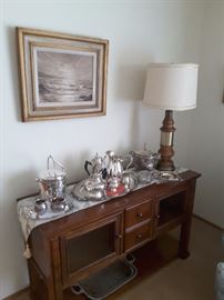 Legends Console with large collection of silver-plated items, ice buckets, serving trays, tea and coffee serving pieces, sugar and creamers, small dishds, beautiful wood lamp base and decorative runner gorgeous Ocean Oil Painting.
