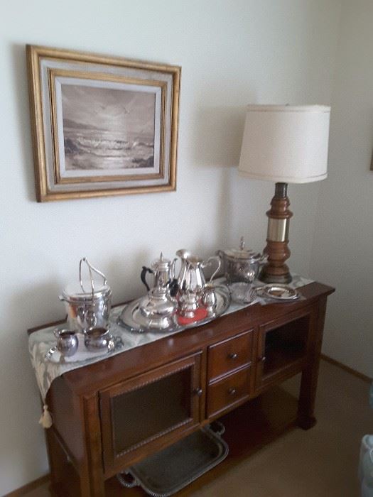 Legends Console with large collection of silver-plated items, ice buckets, serving trays, tea and coffee serving pieces, sugar and creamers, small dishds, beautiful wood lamp base and decorative runner gorgeous Ocean Oil Painting.