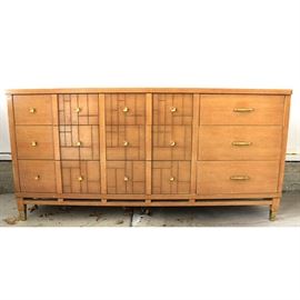 Mid Century Modern Lowboy Dresser: A Mid Century Modern-style lowboy dresser made in the 1950s, The dresser features a a wooden rectangular frame with an oak finish, nine drawers that create a two tone front with three having an etched style and three having part plane and part etched style and blending gold tone modern style pulls with round knobs. The dresser has a modern style with a clean line design and a fretwork style apron with tapered block style feet with gold tone covers.
