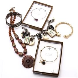 Silver and Gold Tone Stone Jewelry Featuring a Selro Bracelet: A collection of silver tone and gold tone stone jewelry featuring a Selro bracelet. This collection features a soapstone toggle clasp necklace and four silver and gold tone bracelets of which include a story-telling bangle, an elephant cuff, two base metal bangles with dangling druzy stones, and a silver tone Selro bracelet. The Selro bracelet features a snap latch clasp connected to a decorative chain with dangling bone and red glass charms, molded black plastic face charms, and two mahjong-inspired tile charms with “Selro Corp” imprinted upon the clasp.