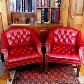 Tufted Leather Office Chairs: A pair of leather office chairs. The chairs feature rolled arms, are upholstered in tufted red leather, and have nailhead trim details. The chairs sit on straight oak legs that are finished in casters.