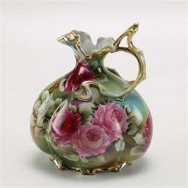 Vintage Nippon Hand-Painted Pitcher: A vintage Nippon pitcher in a floral motif. This picture features an organic free flow style mouth and pour spout with an elaborate single loop, single rest handle in gilt. It has a narrowed neck an bulbous body sitting atop a gilt platform foot with felt coasters. It is decorated in a high gloss finish with images of roses against a green and blue background, and is marked “Nippon Hand Painted” along the underside.