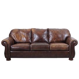 Thomasville Brown Leather Sofa With Ottoman: A Thomasville brown leather sofa with ottoman. This piece has three back cushions attached by zipper, and three seat cushions attached by Velcro. The rolled arms and bottom of the seat front have nail head trim. The sofa sits on bun feet. It is tagged “Thomasville”. A matching ottoman is included. For a coordinating piece, see item 17LOU013-001.