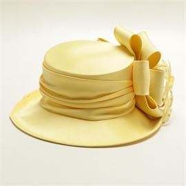 Whitall & Shon Women's Yellow Derby Hat: A Whitall & Shon women’s yellow derby hat. This hat is designed in polyester and features a medium brim with a ruched band and statement bow with feathers and sequin accents. The authentic “Whittall & Shon” trademark label is displayed inside the interior lining. This hat comes complete with a green hat box labeled “Keeneland”.