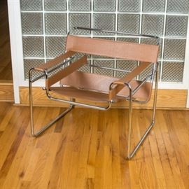 Bauhaus "Wassily" Steel And Leather Armchair After Marcel Breuer: A Bauhaus design “Wassily” chrome plated steel and leather tubular armchair after Marcel Breuer. The chair features a low rectangular bent tube frame, thick wide strips of tan leather stretched to provide back support and create rests for the arms, and a single large piece used for the seat. A label on the underside reads “Made In Italy”. Companion 17BAL014-025.