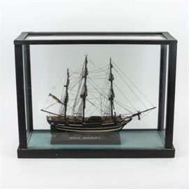 Wooden Model of H.M.S. Bounty Ship and Display Case: A wooden model of H.M.S. " Bounty" in display case. This wooden replica of the ill-fated H.M.S. Bounty is mounted on a wooden base inscribed with the ship’s name and sits in a black wood and glass display case.