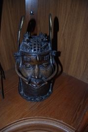 
Africa, Nigeria, Edo, Court of Benin, ca. mid 19th to early 20th century CE. An impressive bronze cast "choker" head depicting an Oba (king of Benin) wearing a netlike coral cap with aliform flaps and a high coral beaded choker. The elaborate coral choker and aliform coral headdress are significant royal court attributes that identify this as the head of an Oba. Size: 7" in diameter x 13.75" H (17.8 cm x 34.9 cm)
The opening at the top of the head was created for elephant teeth or a sculpted elephant tusk carved to depict the many accomplishments of the king. The visage is nicely rendered with thick-lidded open eyes, six decorative scars above them, a straight nose, slightly parted lips, and a row of circular motifs across the cheeks. A piece like this would have been commissioned by a newly enthroned oba to stand on an altar and commemorate a previous oba, most likely his father. The oba of Benin rules via divine right and serves as the being through which his deified royal ancestor