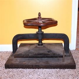 Vintage Book Press: A vintage book press. This press is made of cast iron with a red metallic wheel to the top. It has a flat press and base.