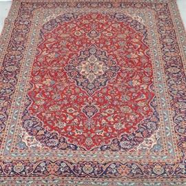Large Handwoven Tabriz Style Rug: A large handwoven Tabriz style wool carpet in blues, reds, greens and cream. The carpet has a vibrant carnelian red field, having an overall pattern of indigo, green, and ivory floral sprays in arabesque patterns throughout. The rug has a Rhodian border with lotus blossoms and poppies on an indigo ground. The rug is hand knotted. Unmarked.