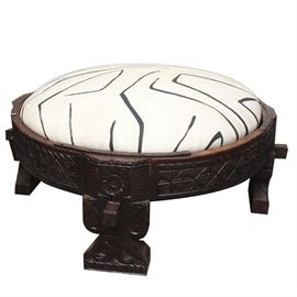 Large Carved Wood and Kelly Wearstler Graffito Upholstered Ottoman: A large and ornately carved wooden ottoman. This round ottoman has four chunky square feet with a scroll and floral motif. The feet support a wide circular frame that has a carved geometric design and hammered metal details on the edge. The ottoman has a rounded pillow center that has been upholstered in a cream and black graphic fabric by Kelly Wearstler called “Graffito”.