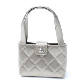 Chanel Silver Satin Evening Bag: A small satin quilted clutch by Chanel. This light beige colored bag has an open top secured by a small fold over tab with a silver toned metal Chanel twist clasp. It has two short carrying straps. The interior has one small zippered pocket with a Chanel-branded zipper pull. The bag has a serial sticker and plastic identification card present. The serial number is 6160705. The bag comes with a black branded duster bag.