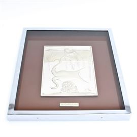 Framed Sculpture in Silver After Salvador Dalí's "Cisne": A silver sculpture after surrealist artist Salvador Dalí (1904-1989) work titled Cisne. This bas relief work is rendered in sterling silver and depicts a female figure seated next to a swan in water. The sculpture is signed to the lower right corner. The work has been framed behind glass in a silver metal shadow box frame that also has a brown tinted glass backside. Within the frame is also a metal marker reading, “Cisne por Salvador Dalí”. On the reverse is a certificate of “uniqueness and authenticity” detailing this work as specimen number 527 / 1500.