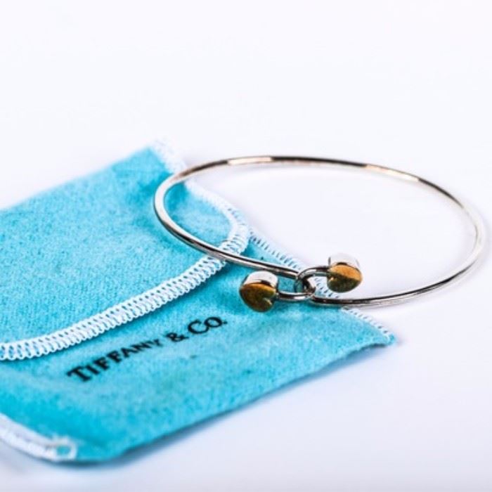 Tiffany & Co. Sterling Silver and 18K Yellow Gold Double Heart Bangle: A Tiffany & Co. sterling silver bangle featuring two interlocking hearts that are accented by an 18K yellow gold overlay. The hearts are both marked “T&Co. 925 750 © 1990” to the verso. This bangle has a total approximate weight of 0.281 ozt and presents in a Tiffany dust bag.