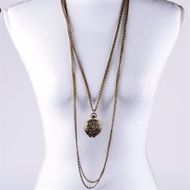 Vintage Signed Goldette Layered Locket Necklace: A vintage Goldette layered locket necklace with a gold plated finish. The round locket features heavily carved floral scrollwork to the exterior and opens with a sprung hinge. It hangs on two snail chains layered with two link chains that are accented by a decorative foldover clasp marked “Goldette N.Y.”. This necklace measures approximately 32" long.