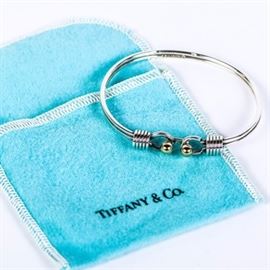 Tiffany & Co. Sterling Silver and 18K Yellow Gold Bangle: A Tiffany &Co. sterling silver bangle featuring two 18K yellow gold beads to the front with an open circle and coiled end caps to each end. The interior is marked “Tiffany & Co. 925, 750” and it has a total approximate weight of 0.474 ozt/9.49 dwt. Presents in a Tiffany dust bag.