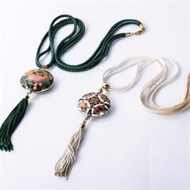 Pair of Cloisonne Necklaces: A pair of cloisonne pendant necklaces. Each pendant features a circular pendant heavily decorated with cloisonne detail, one featuring a butterfly and the other featuring a lotus flower motif. One hangs on a braided green cord with a green tassel and the other hangs on a braided white cord with a white tassel. The items are unmarked and feature base metal barrel clasps.