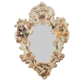 Rococo Style Plaster Wall Mirror: A Rococo style plaster cartouche wall mirror. This ornate piece has a cream plaster frame featuring a man and woman figure along its sides, a cherub at the bottom, and small flowers throughout it. The mirror is marked “Finesse Originals”.