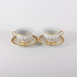 Meissen Porcelain Cups with Saucers: A pair of Meissen porcelain teacups with saucers. Each piece features a white porcelain body with ornate gold-tone embellishments throughout. They are marked with the Meissen double swords on the underside.