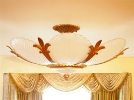 Lot 23: A French art deco style glass and patinated metal semi-flush ceiling fixture. Fleur de lis type brackets with gilt patina alternating with eight frosted glass leaf-form panels, centered by a finial. Good condition. 32" diameter, 15" high. ESTIMATE $600-800