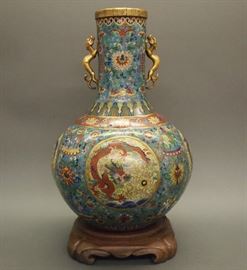 Lot 15: A Chinese gilt bronze and cloisonne vase. Cylindrical neck flanked by mounted dragons above a bulbous base with four circular reserves about, each is slightly raised and encloses a dragon in pursuit of a flaming pearl, on a bright blue ground accented with elephants and recumbent rams among flowers. Qianlong style mark underneath, probably 20th century. Numerous small areas of chipped enamel, including the shoulder, sides, and about the base. 17" high, raised on a separate wood base. ESTIMATE $800-1,200