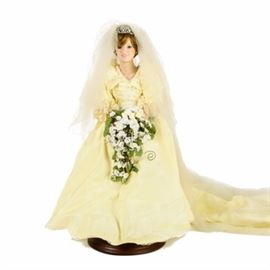 Danbury Mint Porcelain "The Princess Diana Bride Doll": A Danbury Mint The Princess Diana Bride porcelain doll. The doll features bisque hands and a head with brown hair and blue eyes. She wears a yellow and cream dress accented with cream colored gemstones around the waist. A pair of yellow heels, a long flowing lace veil and a white and green bouquet complete her outfit. She has a soft body. The back of the doll’s neck reads "The Princess Diana Bride Doll with a “D” over an “M” for the Danbury Mint.