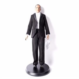 Danbury Mint "Barack Obama" Porcelain Doll: A Danbury Mint porcelain Barack Obama doll. The doll is wearing a black tuxedo with a white shirt, and an American flag pin on his lapel. This piece includes an original tag and is marked “SD-949” to the back of the neck and also includes a display stand.