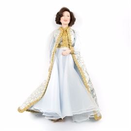 Danbury Mint "Jacqueline Kennedy" Porcelain Doll: A Danbury Mint Jacqueline Kennedy porcelain doll. The doll features bisque hands and a head with brown hair and brown eyes. She wears a blue and white dress with gold-tone lace trim to the edges of the overcoat and gold heals to complete her outfit. She has a soft body. The back of the doll is marked with a “D” over an “M” and “© MBI.”
