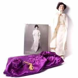 Danbury Mint "Jaqueline Kennedy" Porcelain Doll and Book: A Danbury Mint Jacqueline Kennedy porcelain doll and book. The doll features bisque hands and a head with brunette hair and brown eyes. She wears a replica of the off-white peau d’ange sleeveless dress with chiffon overlay designed by Oleg Cassini, worn to President John F. Kennedy’s inauguration in 1960. An off-white cape that closes at the neck, faux pearl drop earrings and opera length gloves complete the outfit. A dark wood stand and purple satin bag is included. The hardcover exhibition catalog from the 2001 Metropolitan Museum of Art, New York, exhibition titled Jacqueline Kennedy: The White House Years, Selections from the John F. Kennedy Library and Museum by Hamish Bowles with a forward by Arthur M. Schlesinger, Jr. and Rachel Lambert Mellon is included.