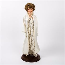 Danbury Mint "Nancy Reagan" Porcelain Doll: A Danbury Mint Nancy Reagan porcelain doll. The doll features bisque hands and a head with brunette hair and brown eyes. She wears a replica of the white, one shouldered, beaded and lace, James Galanos. sheath gown, worn to President Ronald W. Reagan’s first inauguration in 1981. A white sleeved cape with ruffled collar, opera length gloves, silver-tone earrings and David Evins heels complete the outfit. A dark wood stand is included.