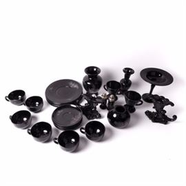 Collection of Black Glass Tableware Including Fenton: A collection of black glass tableware, including hand-painted Fenton pieces. The selection includes two Fenton vases and a rose bowl in black amethyst glass, hand-painted in silver by Diane Gessel, featuring a silver floral motif and marked “Hand Painted by D. Gessel” on the verso. Included are six Fenton black amethyst coffee cups, also hand-painted and marked by Diane Gessel, featuring a silver floral motif with six matching saucers. Six unmarked plates are in the same silver motif are included, with a black milk glass sugar bowl and creamer with a silver floral motif; and a black salt and pepper shaker set with a stacked “V(?)” mark and silver floral embellishment. Also included are two black ceramic candle holders in a scrolled trumpet shape, unmarked, and a tall pedestal black glass compote, unmarked, with a pontil mark to the underside.