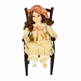 1981 Gorham "Rosamond" Musical Wind Up Porcelain Doll and Vintage Doll Chair: A 1981 Gorham Rosamond musical wind up porcelain doll and vintage doll chair. The doll features a yellow lace gown accented with a pink bow and bonnet. She has brown curly hair and brown eyes, and also a gold tone bracelet with a letter “G” for Gorham. The back of her neck is marked “Gorham 1981” and she included the original wind up key. The doll is also perched on a vintage Chippendale-style chair with an arched slat back that has an upholstered floral seat and is marked “American Classics-Doll Furniture”.