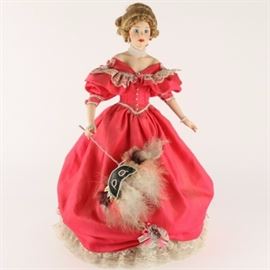 1990 Danbury Mint Red Masquerade Ball Porcelain Doll: A 1990 Danbury Mint red masquerade ball porcelain doll. The doll features bisque hands and a head with blonde hair and blue eyes. She wears a red dress with a lace trimmed, and is holding a masquerade mask with feathers. Red pantalettes, socks and red boots complete her outfit. She has a soft body with a large faux pearl beaded necklace and faux pearl earring strands. The back of the doll features a “d” over “m” mark with “1990 MBI”. She is also equipped with a wooden stand.