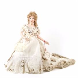 Hamilton "Alexandra" Porcelain Doll: A Hamilton “Alexandra” porcelain doll. The doll features bisque hands and a head with strawberry blonde hair and blue eyes. She wears a white and lace trimmed dress that is accented with a long dress train. The dress also features a plethora of white, pink and purple rosettes with a fringed trim along the train and running up the dress. White pantalettes faux pearl and gold tone neckalce and earring sets with a pair of white heals complete her outfit. She has a soft body. The back of the doll is marked “Alexandra © 1990 The Hamilton Collection” and also has the original tag hanging from her left hand.