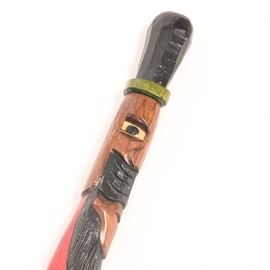 Carved Wooden Cane: A carved wooden cane. This cane is carved similarly to a totem that terminates into a handle. In addition to being carved, the cane is painted in a palette of black, green, and pink. The rest of the cane is stained and the entire cane has a lacquered finish.